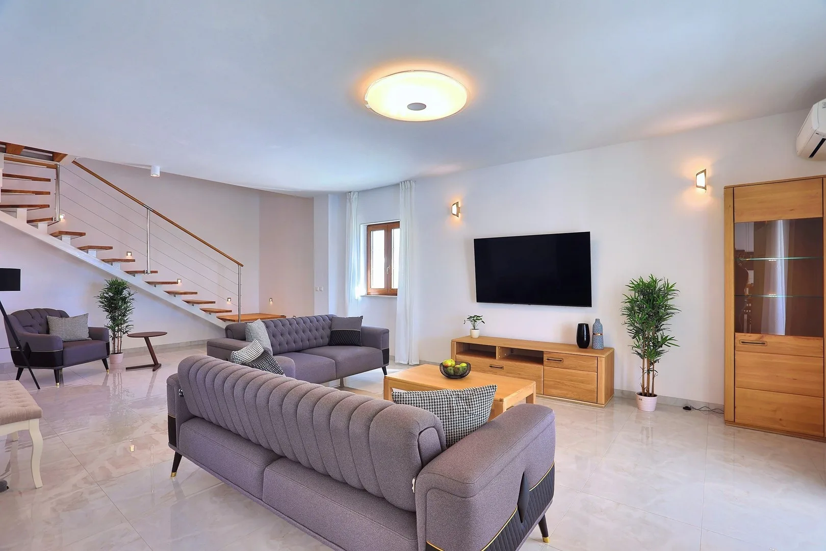 A bright living room with gray seating, a television, and a staircase.