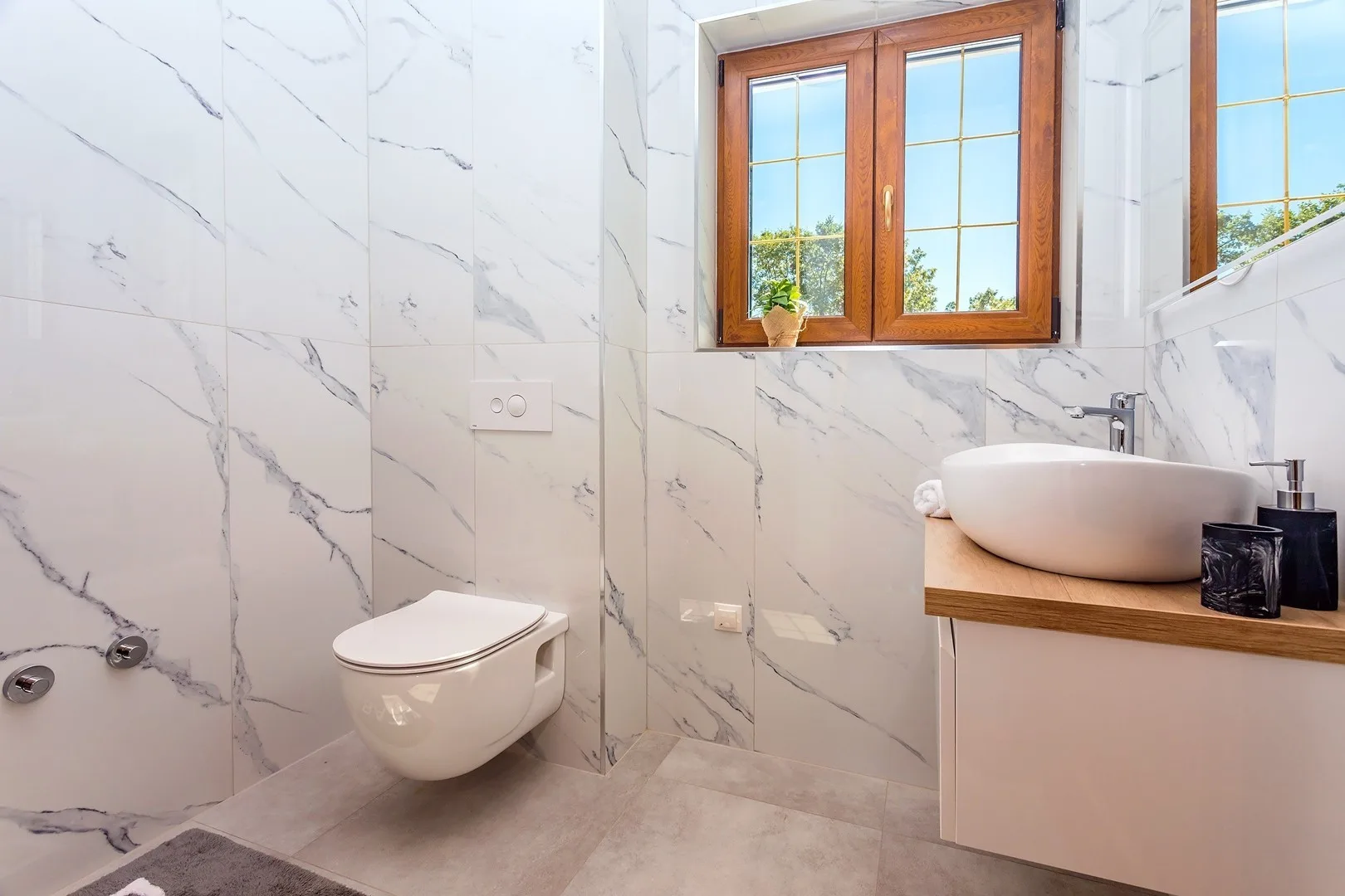 A bright tiled bathroom with a wall-mounted toilet and a basin beneath a window.