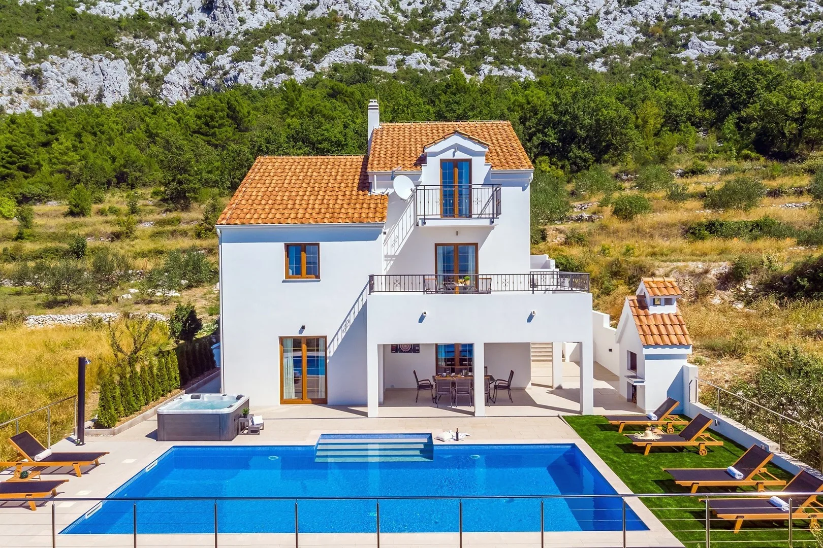A white house with a swimming pool and sun loungers sits among green trees.