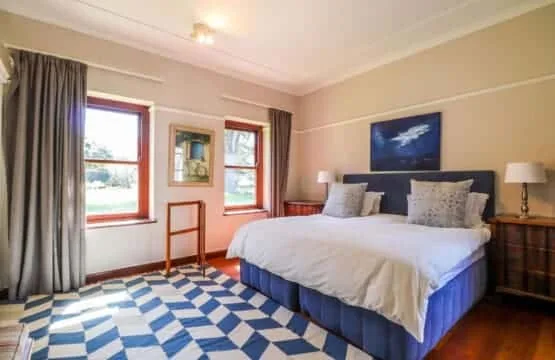 A bedroom with a large bed, blue-and-white checkered rug, and two curtained windows.