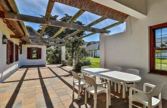 A sunny tiled patio has a long white table and chairs beneath an open timber pergola.
