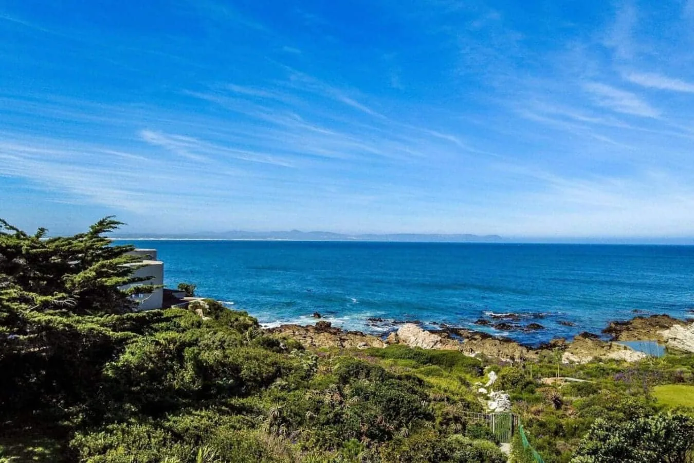 A rocky coastline with dense green shrubs overlooks the blue sea beneath a clear, streaked sky.