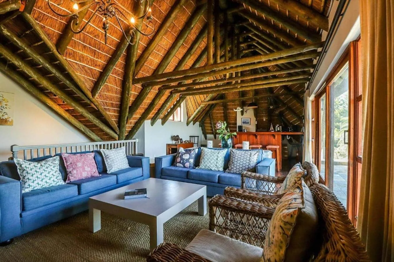A spacious living room has blue sofas, woven chairs, a coffee table, and an exposed timber-and-thatch ceiling.