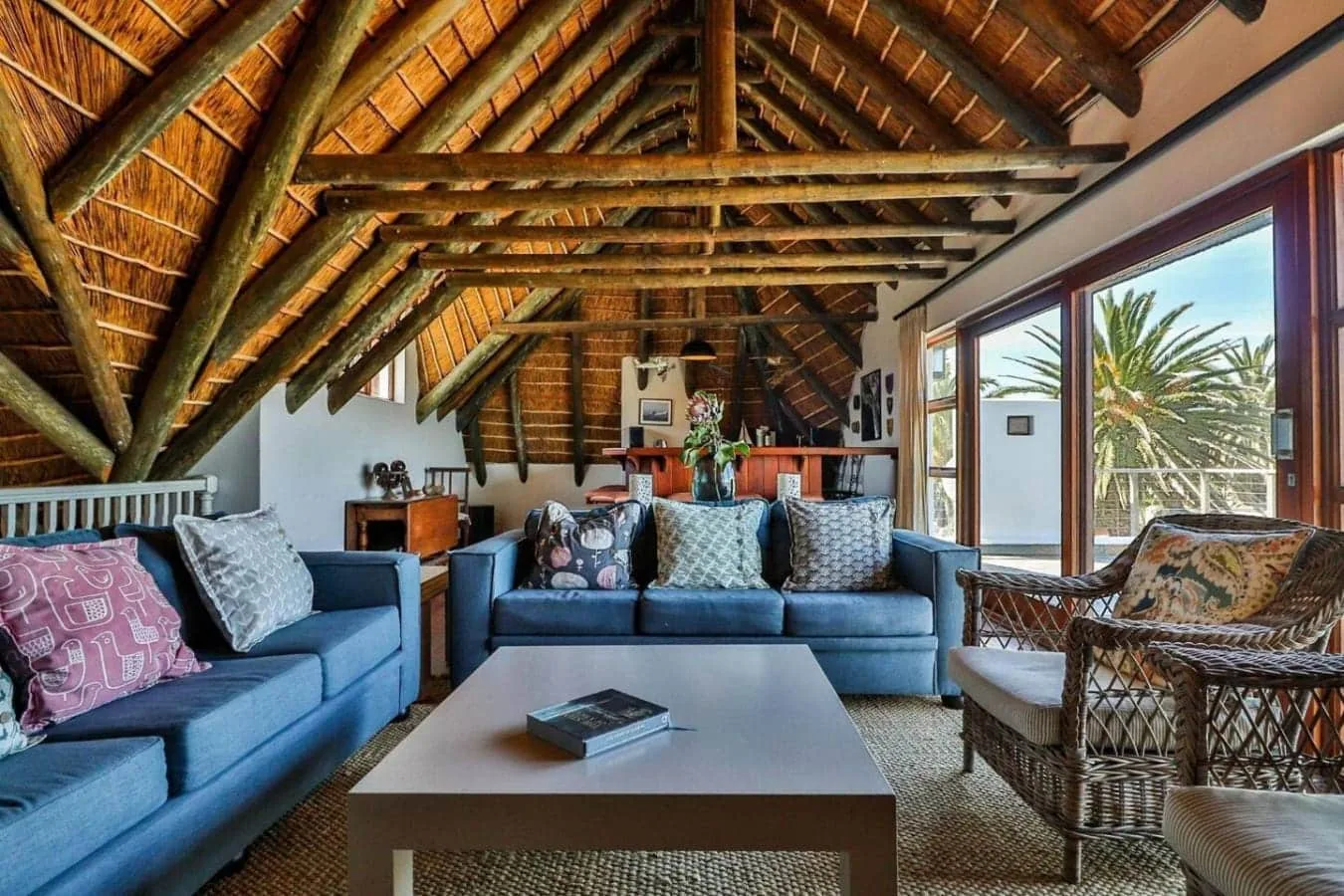 A living room with blue sofas, wicker chairs, and a coffee table beneath an exposed timber roof.