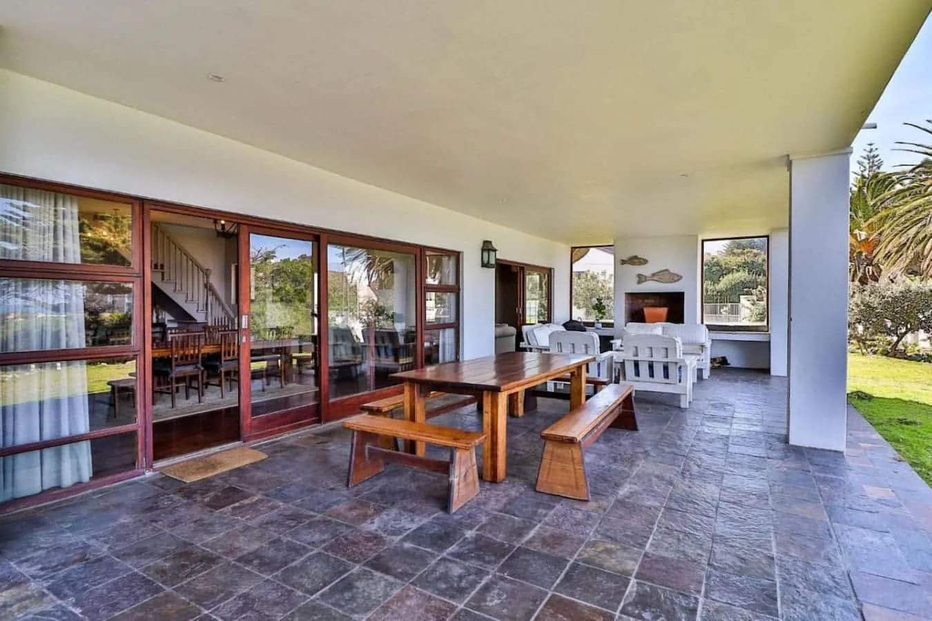 A covered patio has a long wooden picnic table, white seating, and glass doors overlooking the garden.