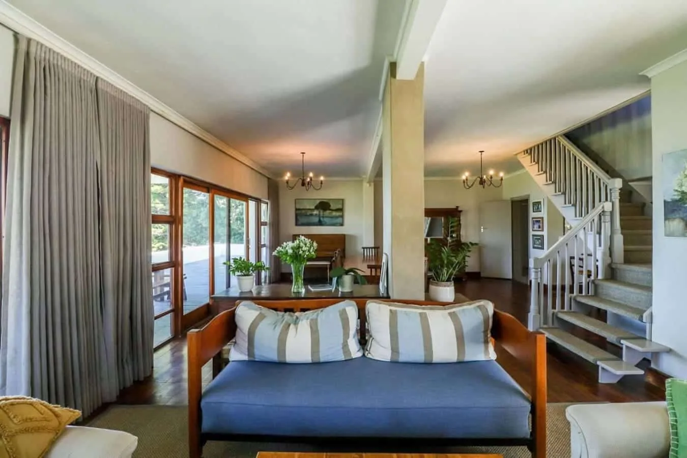 A bright living room has a blue sofa, large glass doors, and a staircase beside a dining area.