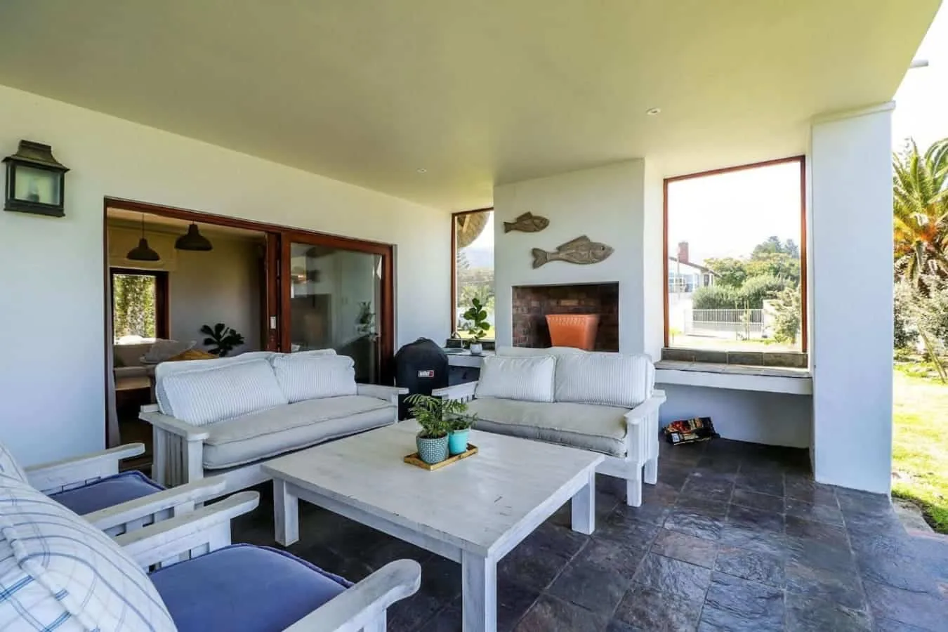 An open-air living area has white sofas, a low coffee table, and a fireplace beside large windows.