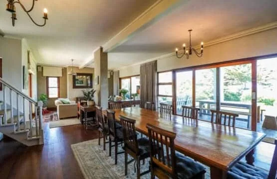 A spacious dining room has a long wooden table, chairs, chandeliers, and large windows.