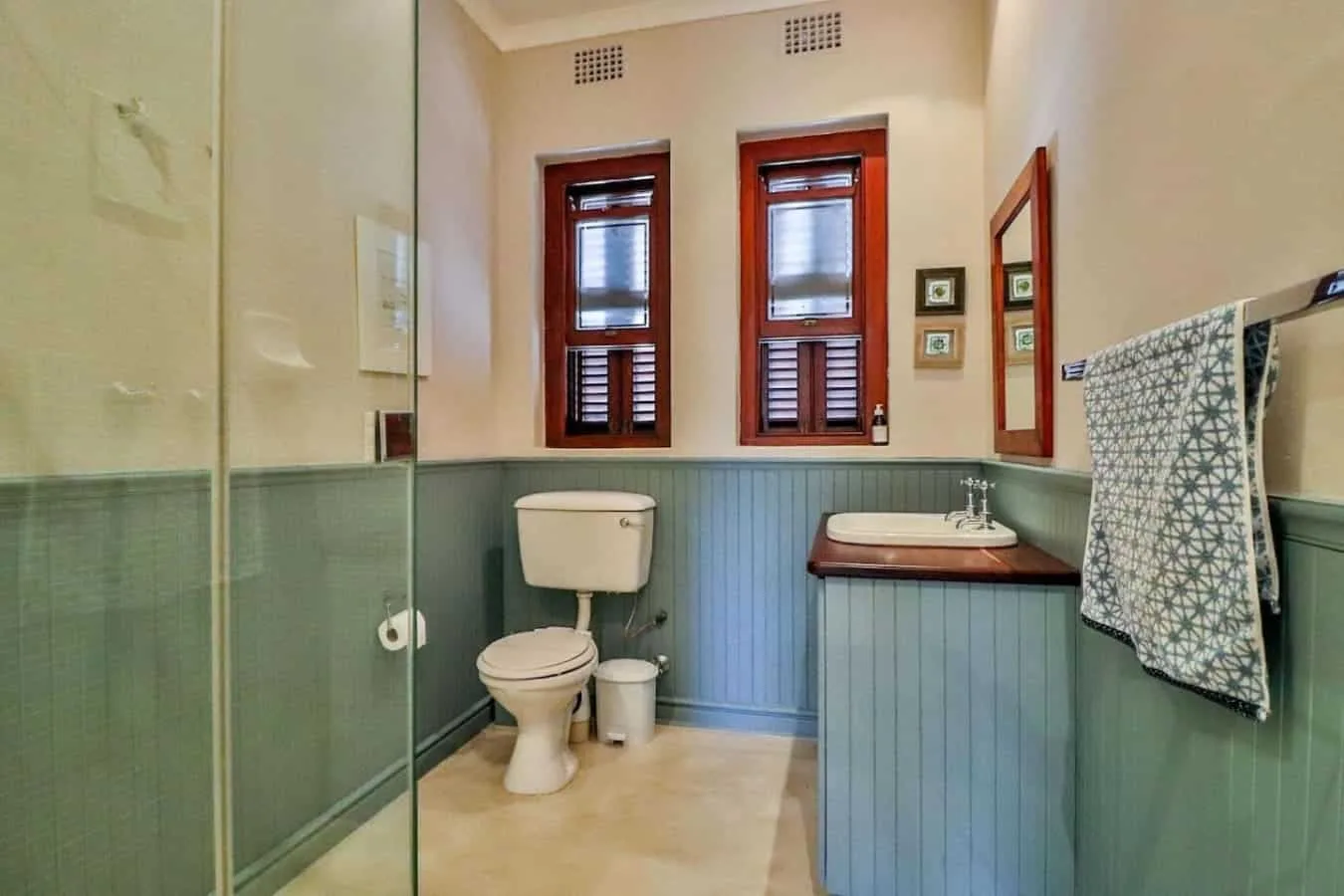 A bathroom with a glass shower enclosure, toilet, and sink beneath two shuttered windows.