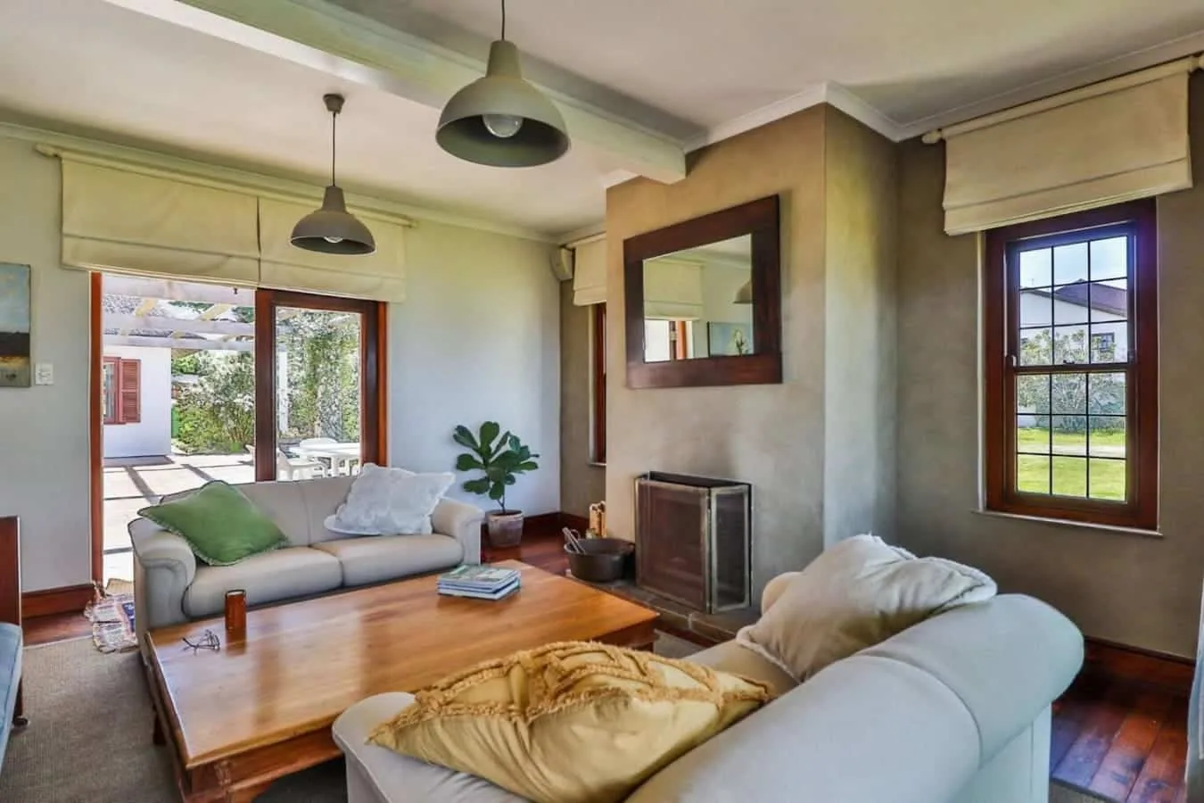 A bright living room has pale sofas, a wooden coffee table, and windows opening onto a garden.
