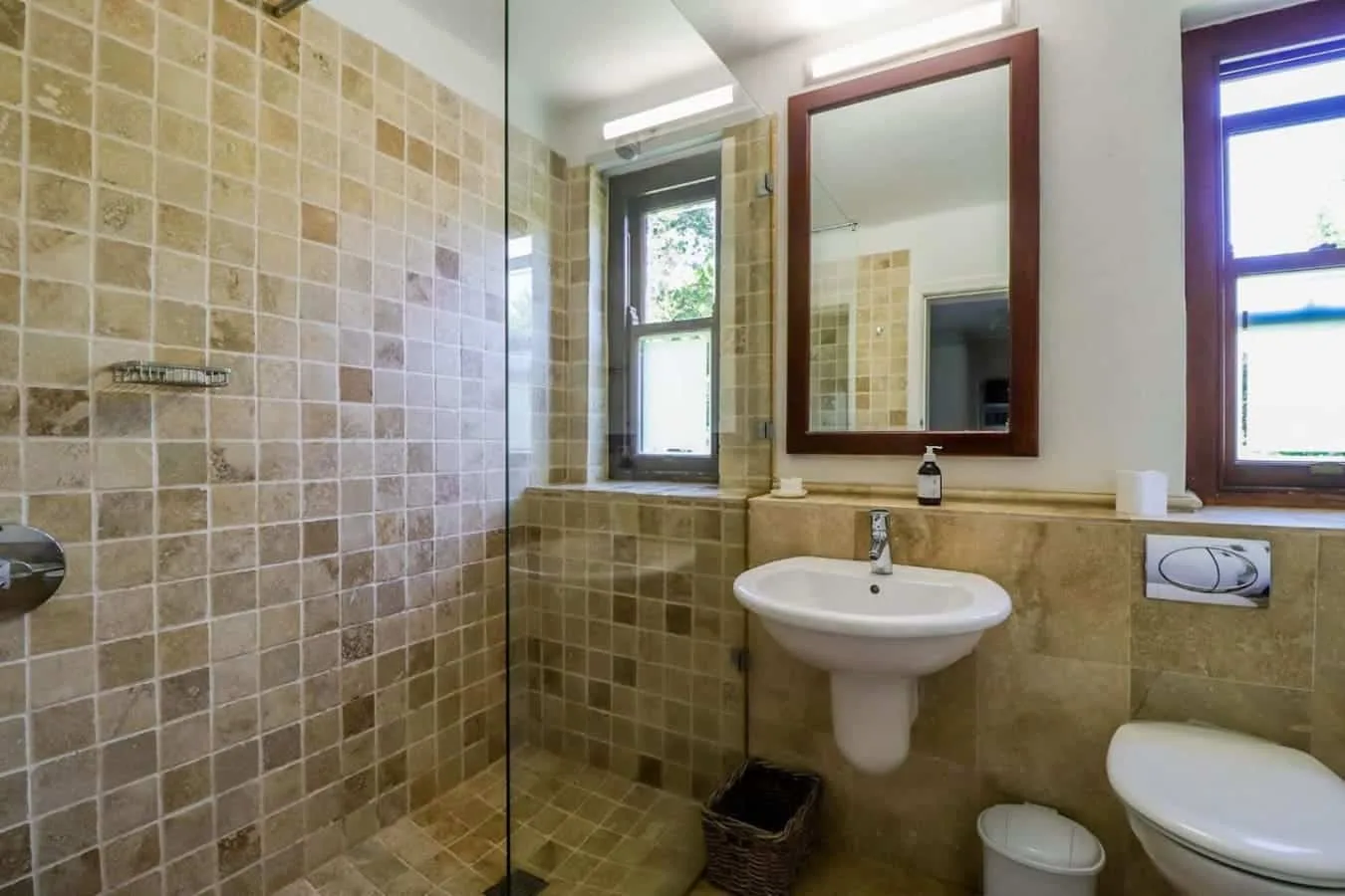 A tiled bathroom has a glass shower enclosure, pedestal sink, mirror, and toilet.