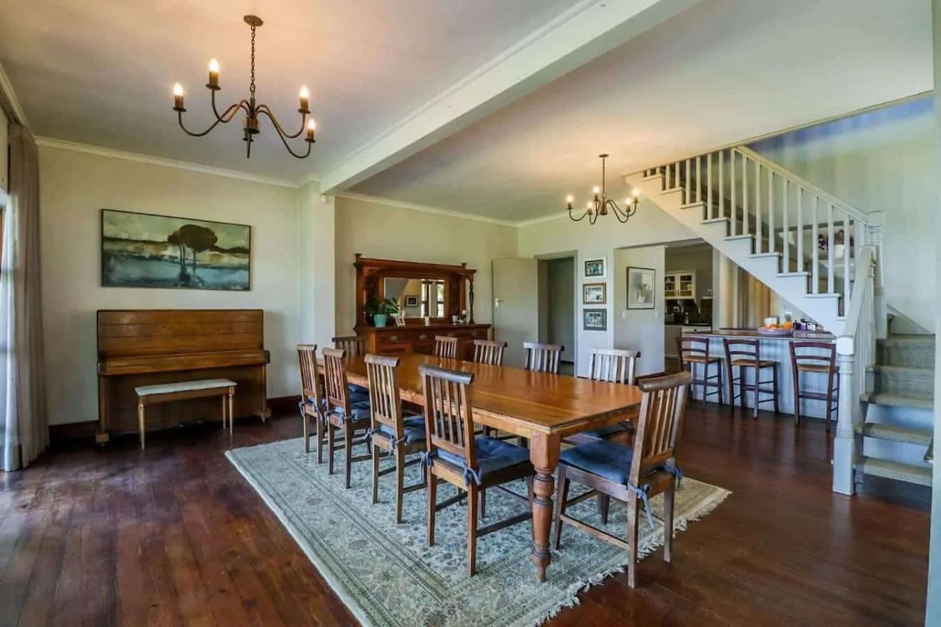 A spacious dining room has a long wooden table, upright piano, chandeliers, and a staircase.