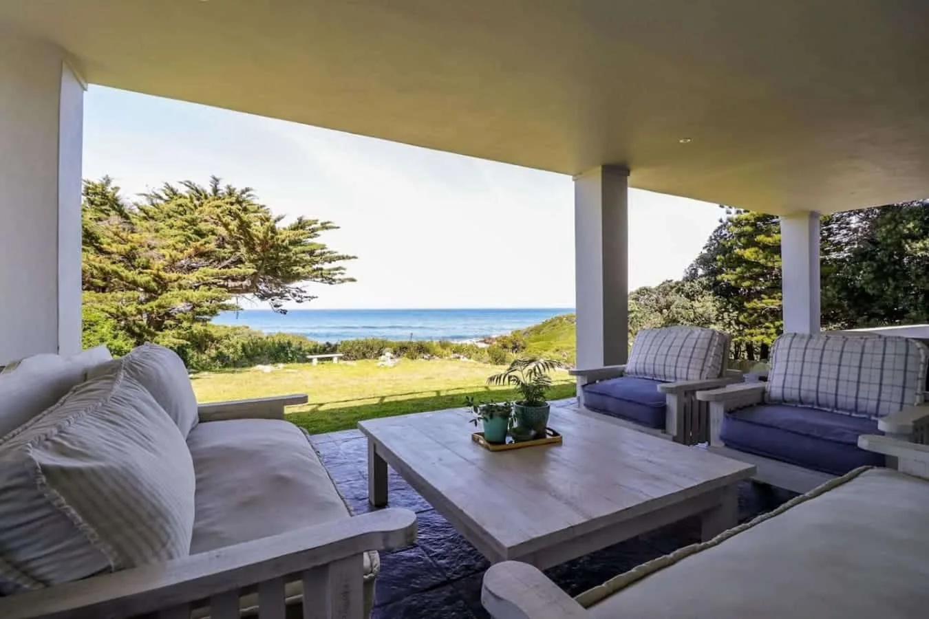 A covered patio overlooks the ocean, with cushioned seating, a low table, and greenery.