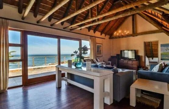 A living room with exposed wooden ceiling beams, gray sofas, and large windows overlooking the ocean.