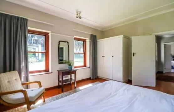 A bright bedroom has a white bed, two tall windows, a wardrobe, and a wooden chair.