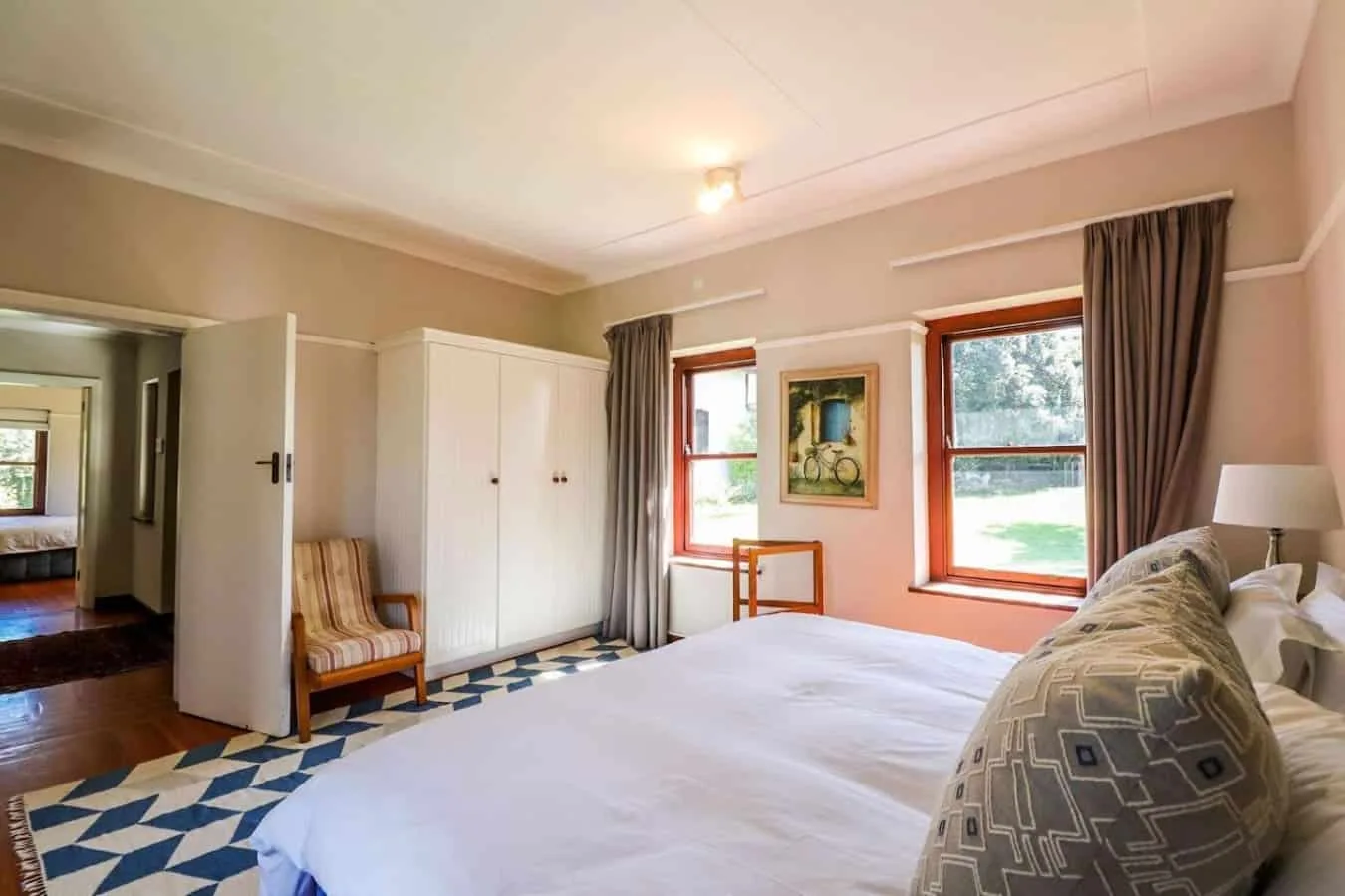 A bright bedroom with a large white bed, two windows, and a wardrobe beside the open doorway.