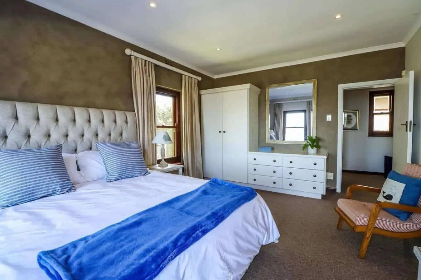 A bedroom with a large bed, white wardrobe and dresser, and a wooden chair beside the doorway.