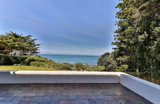Outdoor terrace with a tiled floor and low white wall overlooking the ocean, framed by trees.