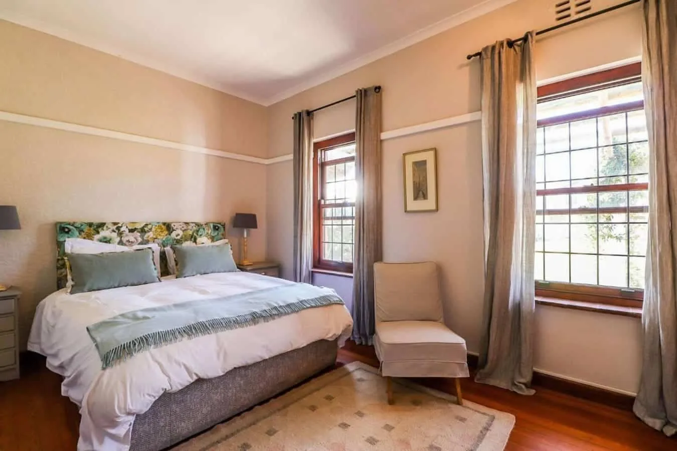 A bedroom with a double bed, patterned headboard, two tall windows, and a light upholstered chair.
