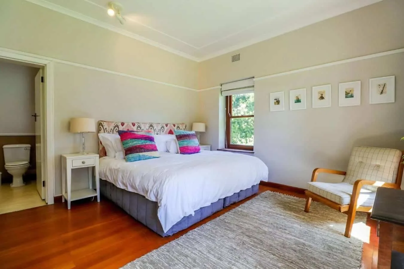 A bright bedroom features a large bed with colorful pillows, a window, and a woven rug.