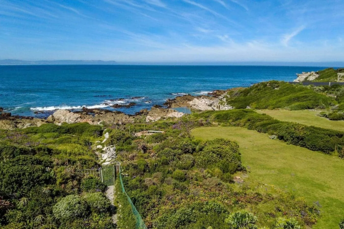 A grassy coastal bluff with low shrubs overlooks rocky shoreline and blue ocean beneath a clear sky.