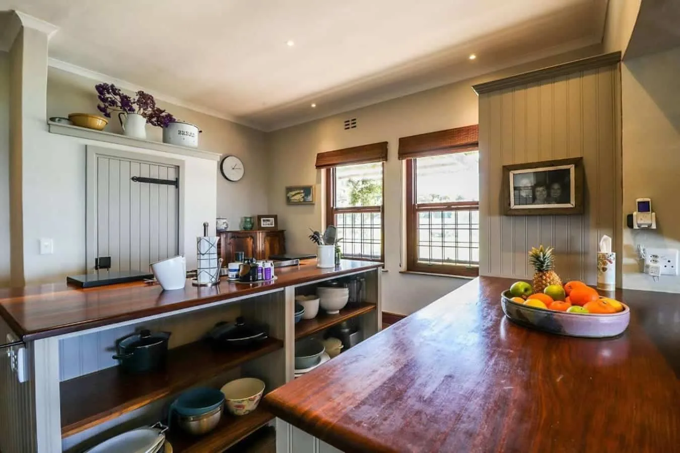A kitchen with long wooden counters, open shelves, two windows, and a bowl of fruit.