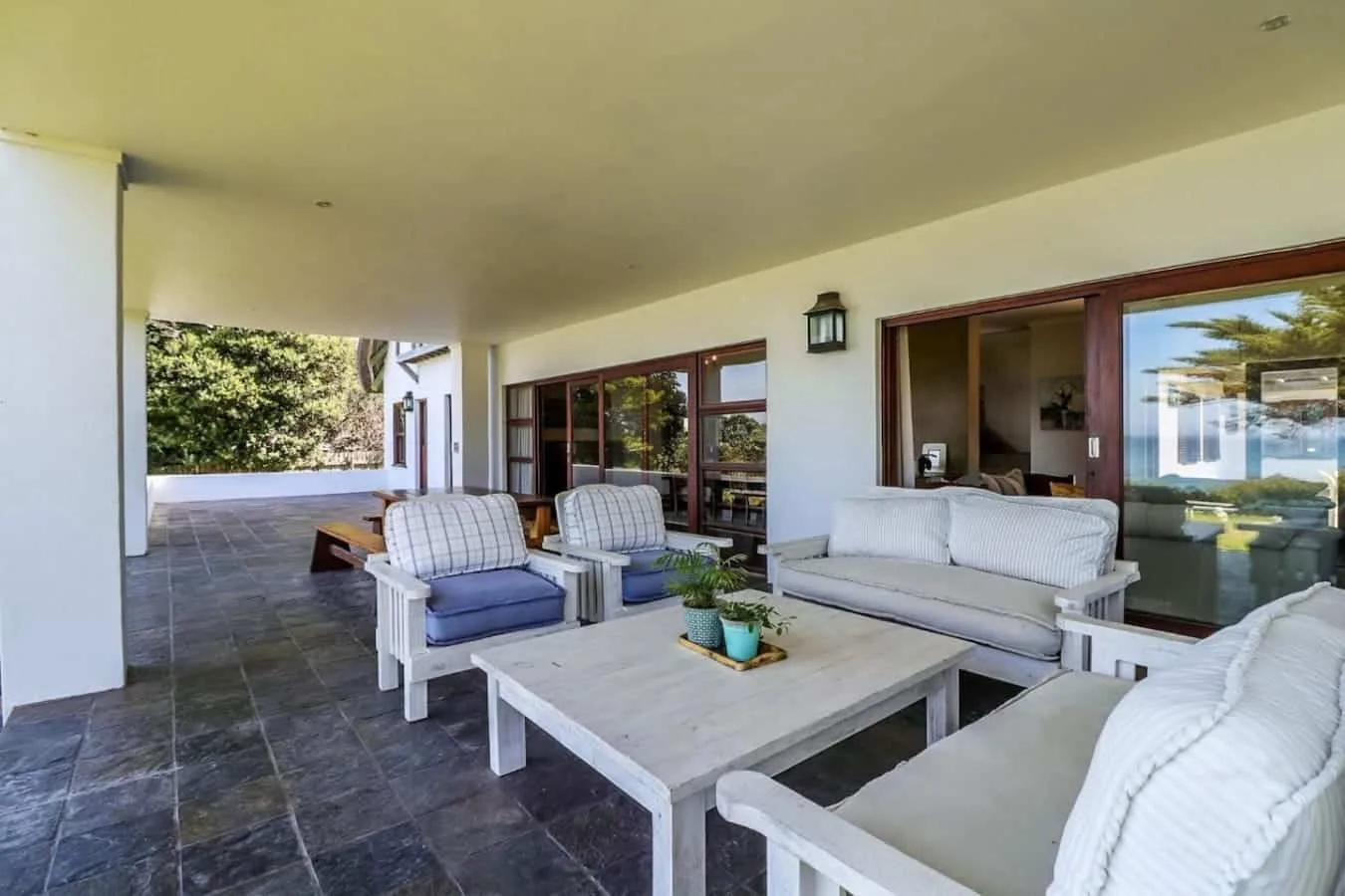 A covered patio has white outdoor sofas and armchairs around a low coffee table beside glass doors.