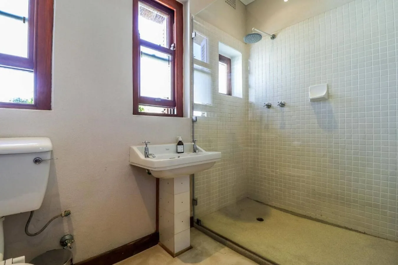 A tiled bathroom has a walk-in shower, pedestal sink, toilet, and two tall windows.
