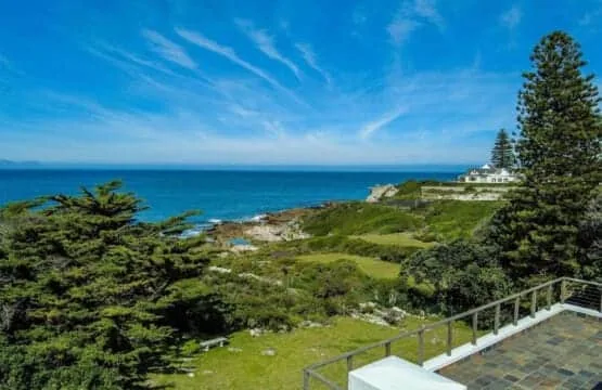 A paved terrace overlooks green coastal grounds, rocky shoreline, and the blue ocean.