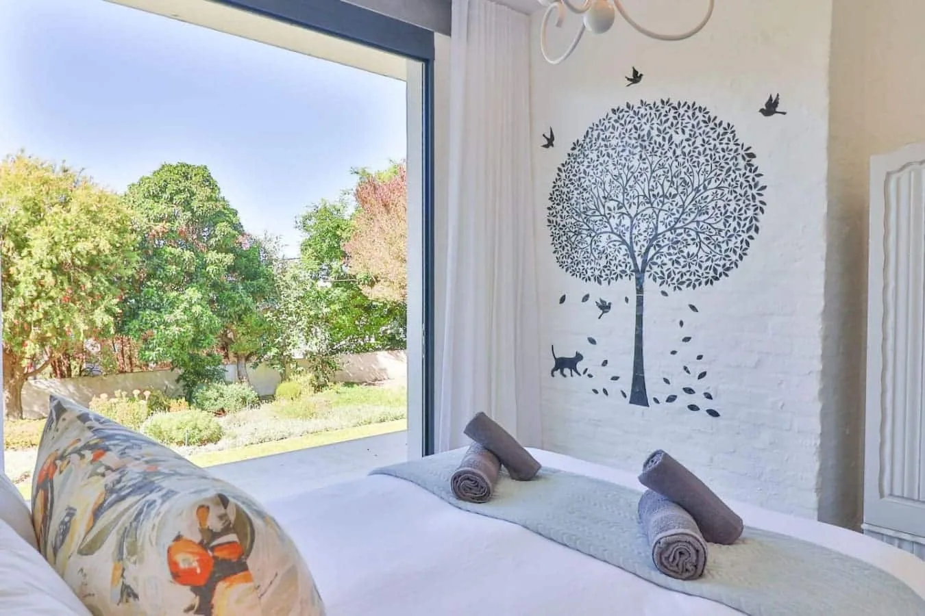 A bedroom with a bed beside a large window overlooking leafy greenery.