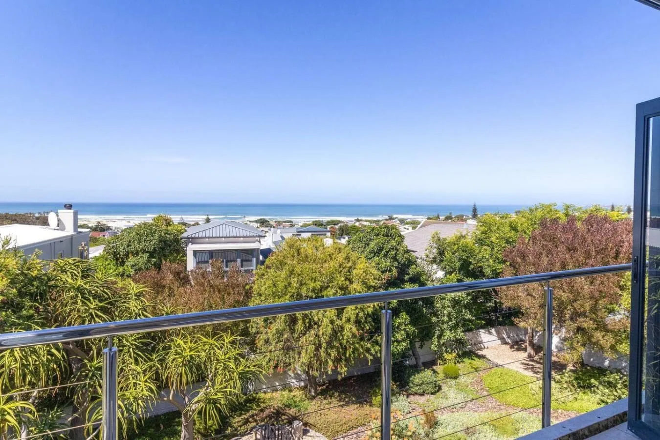 A balcony overlooks leafy gardens and the ocean beneath a clear sky.