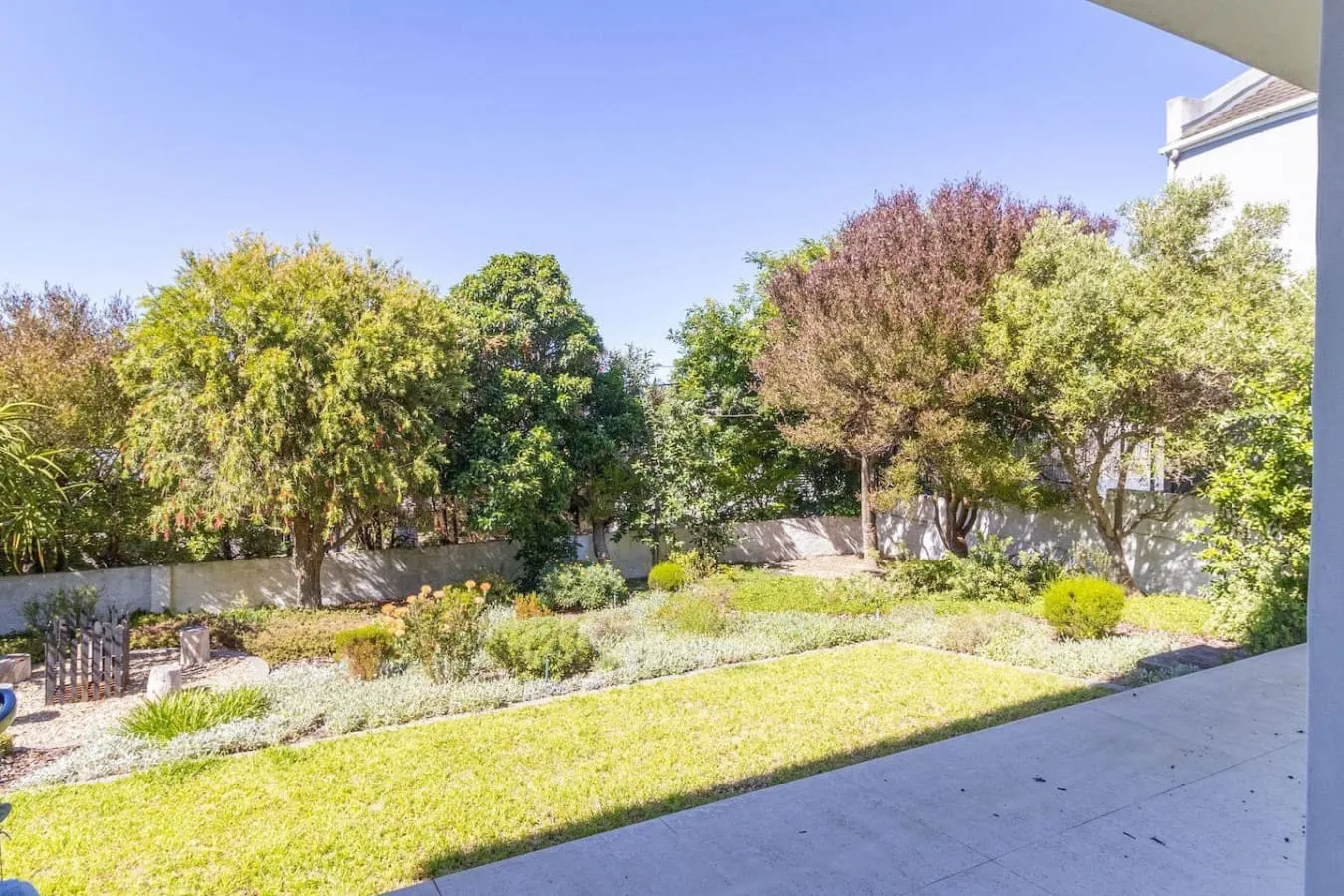 A garden with lawn, low plantings, and mature trees is viewed from a covered patio.