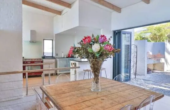 A dining table with a flower arrangement sits beside an open kitchen and a glass doorway.