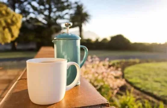 Una taza blanca y una cafetera color turquesa están sobre una mesa al aire libre junto a un jardín.