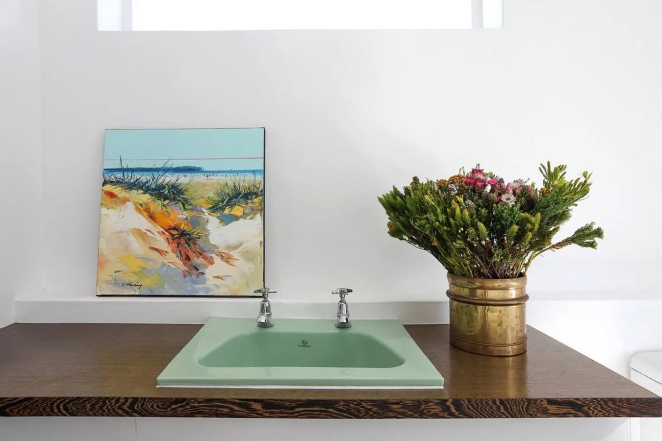 Un lavabo de baño con un fregadero verde, una obra enmarcada y flores en un jarrón.