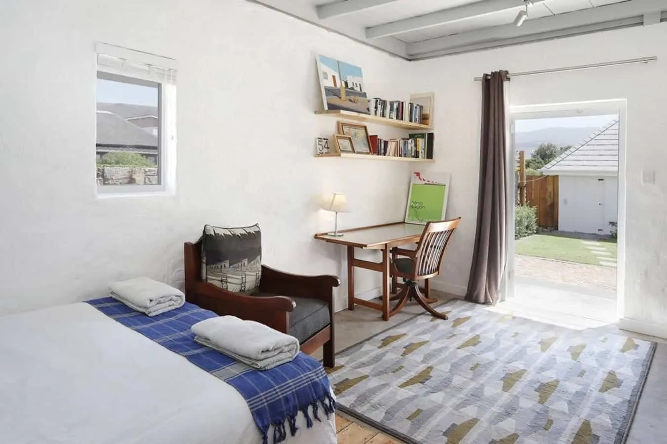 Un dormitorio luminoso tiene una cama, un sillón y un pequeño escritorio junto a la puerta abierta.