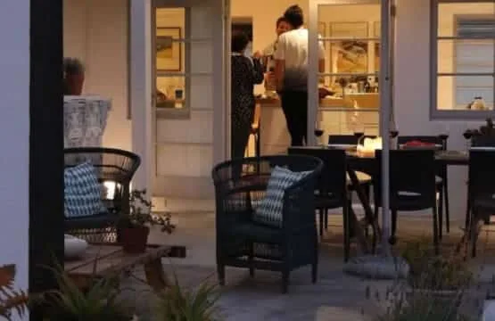 Una zona de estar en el patio da a una habitación iluminada con personas dentro.