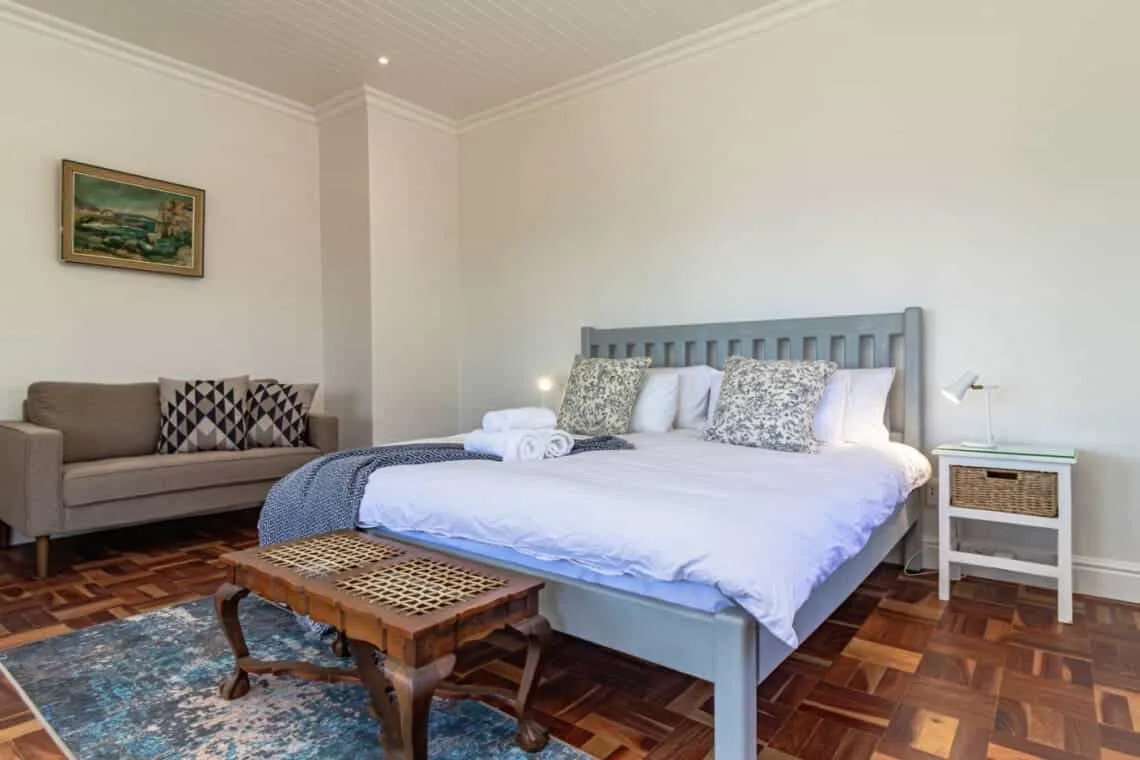 Un dormitorio luminoso cuenta con una cama grande, un sofá con cojines estampados y una pequeña mesa de centro de madera.