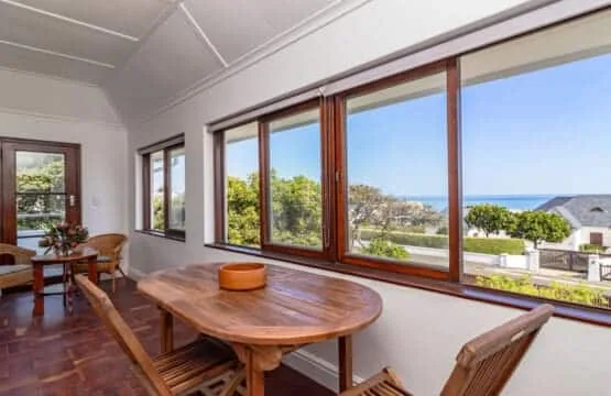 Un comedor luminoso cuenta con una mesa de madera, sillas y grandes ventanales con vistas a árboles y al mar.