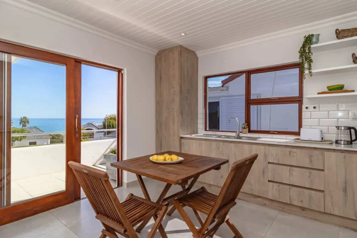 Una luminosa cocina con comedor, mesa de madera, armarios empotrados y ventanas con vistas al mar.