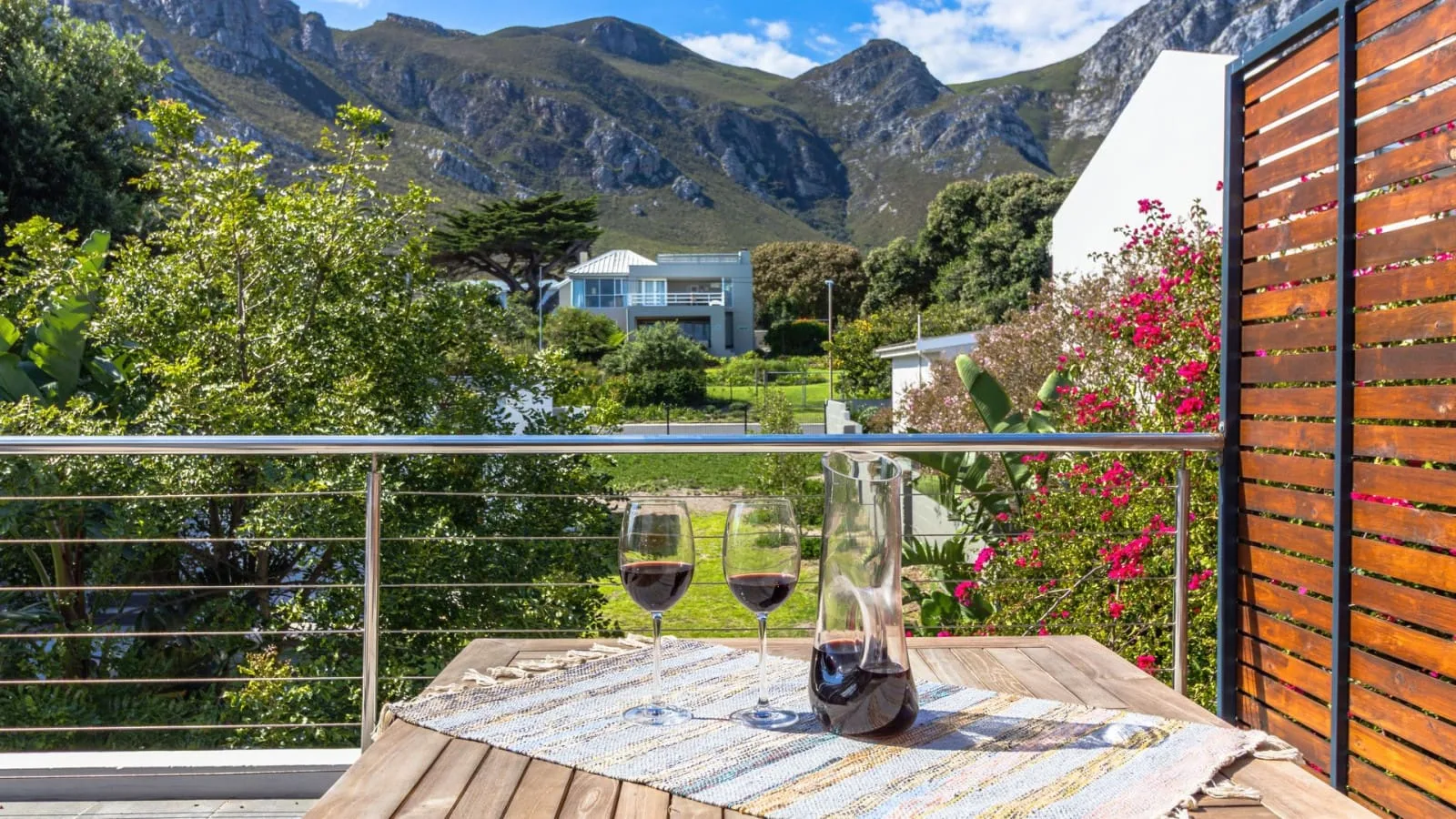 Una terraza de madera da a un jardín frondoso y una cordillera, con copas de vino sobre una mesa.
