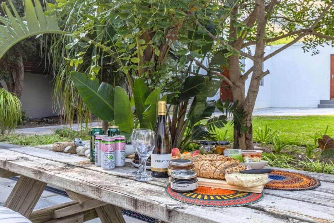 Una mesa de picnic sostiene bebidas, frascos y pan bajo árboles frondosos, junto a un césped.