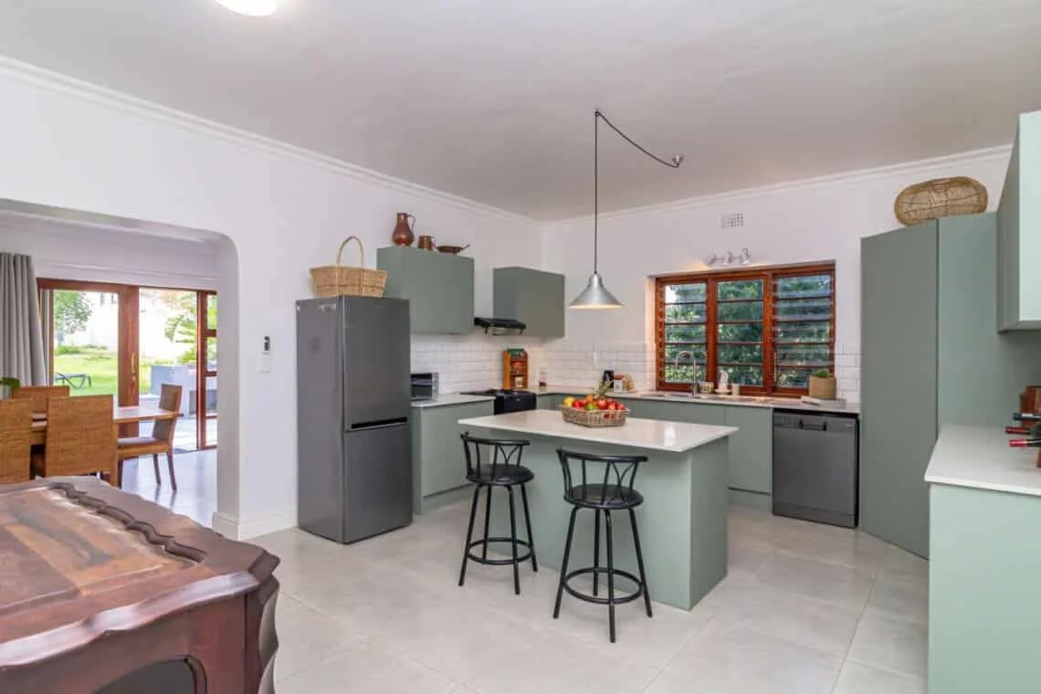 Una cocina luminosa tiene gabinetes grises, una isla central con taburetes y una ventana con marco de madera.