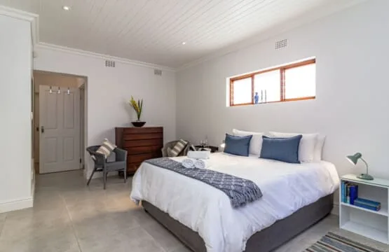 Un dormitorio luminoso tiene una cama grande con almohadas azules, una cómoda de madera y dos sillas.