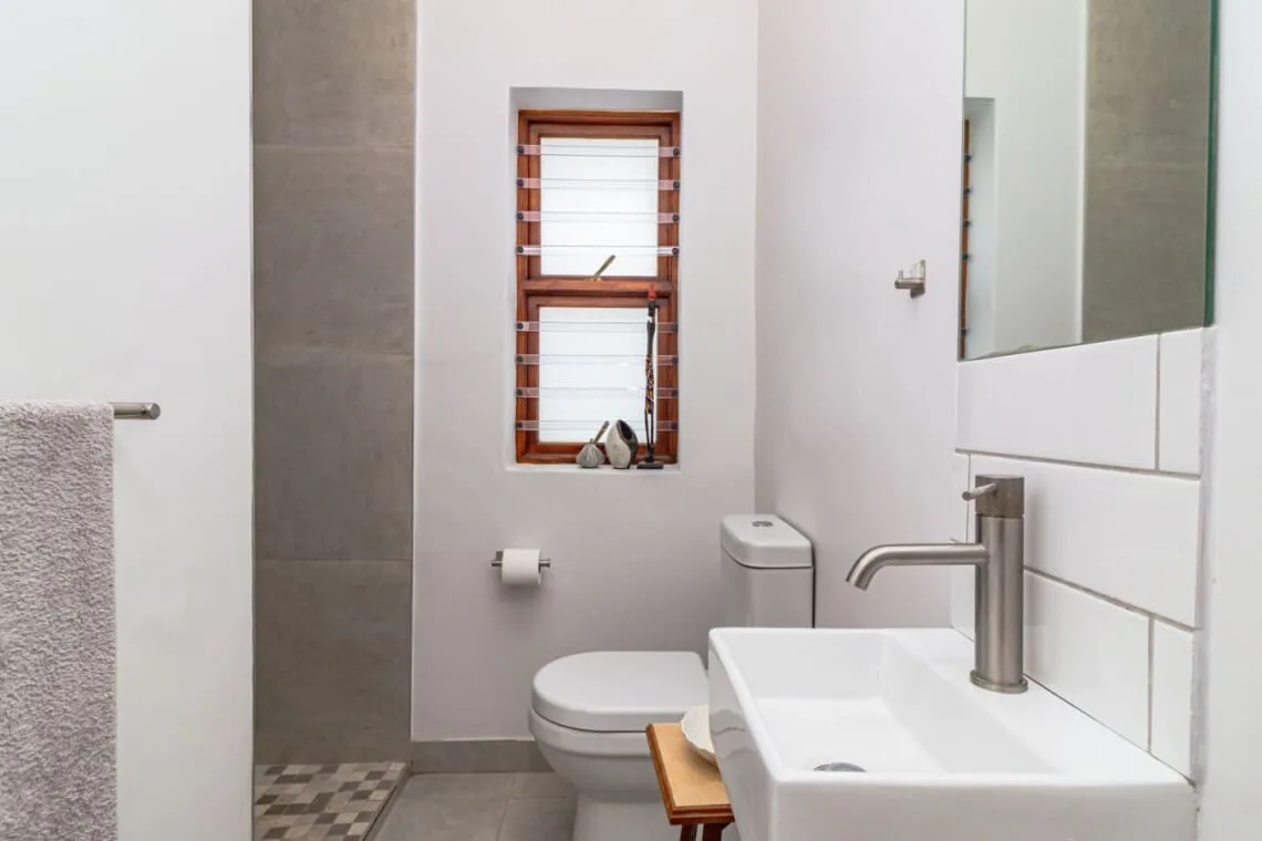 Un baño moderno con inodoro blanco, lavabo rectangular, espejo de pared y una ventana pequeña con persianas.