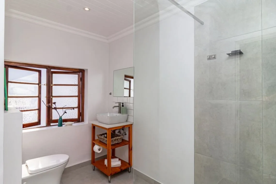 Baño con ducha de mampara de vidrio, lavabo blanco sobre un mueble de madera y dos ventanas.