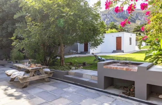 Un patio ajardinado pavimentado tiene una mesa de picnic de madera, árboles frondosos y un banco bajo junto a un edificio blanco.
