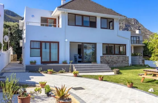 Una casa blanca moderna de dos plantas da a un patio pavimentado y un césped, con plantas en macetas y una mesa de pícnic.