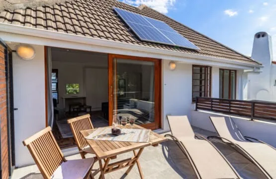 Una terraza soleada tiene sillas de madera, tumbonas y una mesa junto a una puerta de cristal.