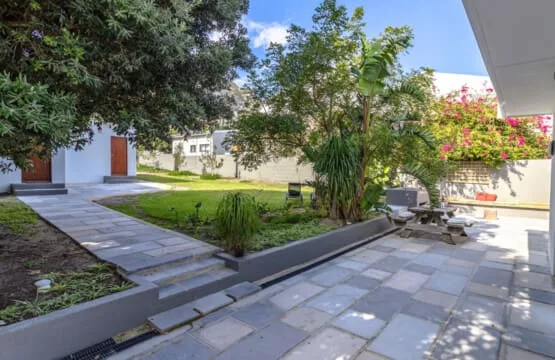 Un sendero pavimentado bordea un pequeño jardín con árboles, arbustos y asientos al aire libre.